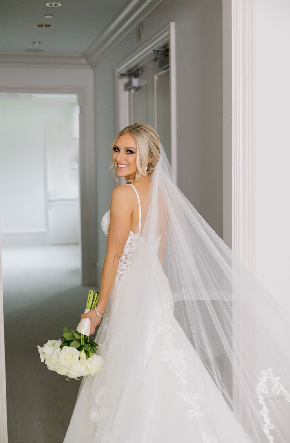 Full-length bridal portrait showing detailed lace gown and flowing veil in bright hallway.