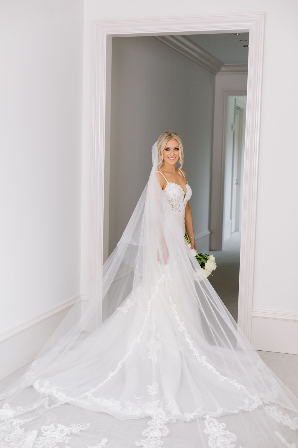 Elegant bride in fitted lace wedding gown with long flowing veil poses in white doorway.