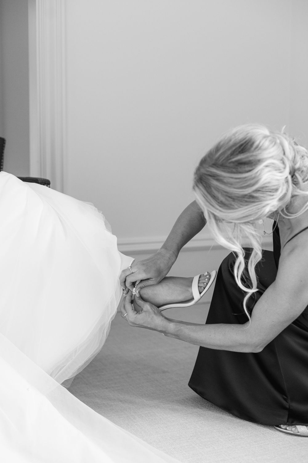 Black and white image of wedding dress preparation with attendant adjusting bridal shoes.
