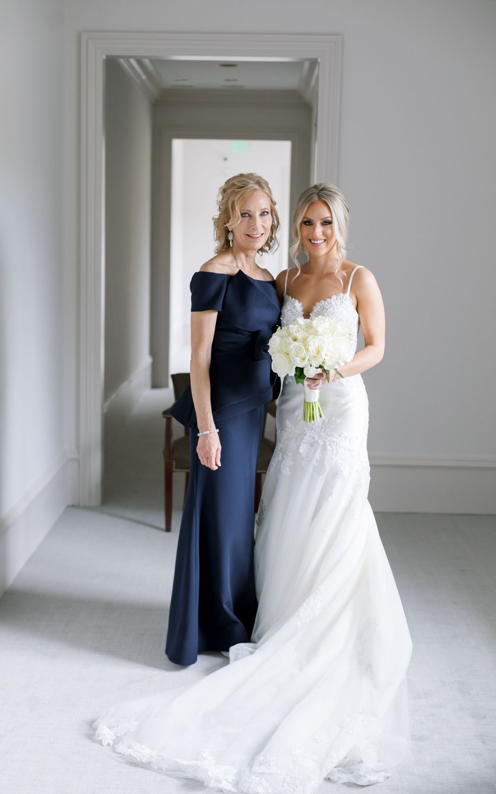 Elegant wedding portrait featuring a navy blue formal gown and white bridal dress with bouquet in minimalist hallway.