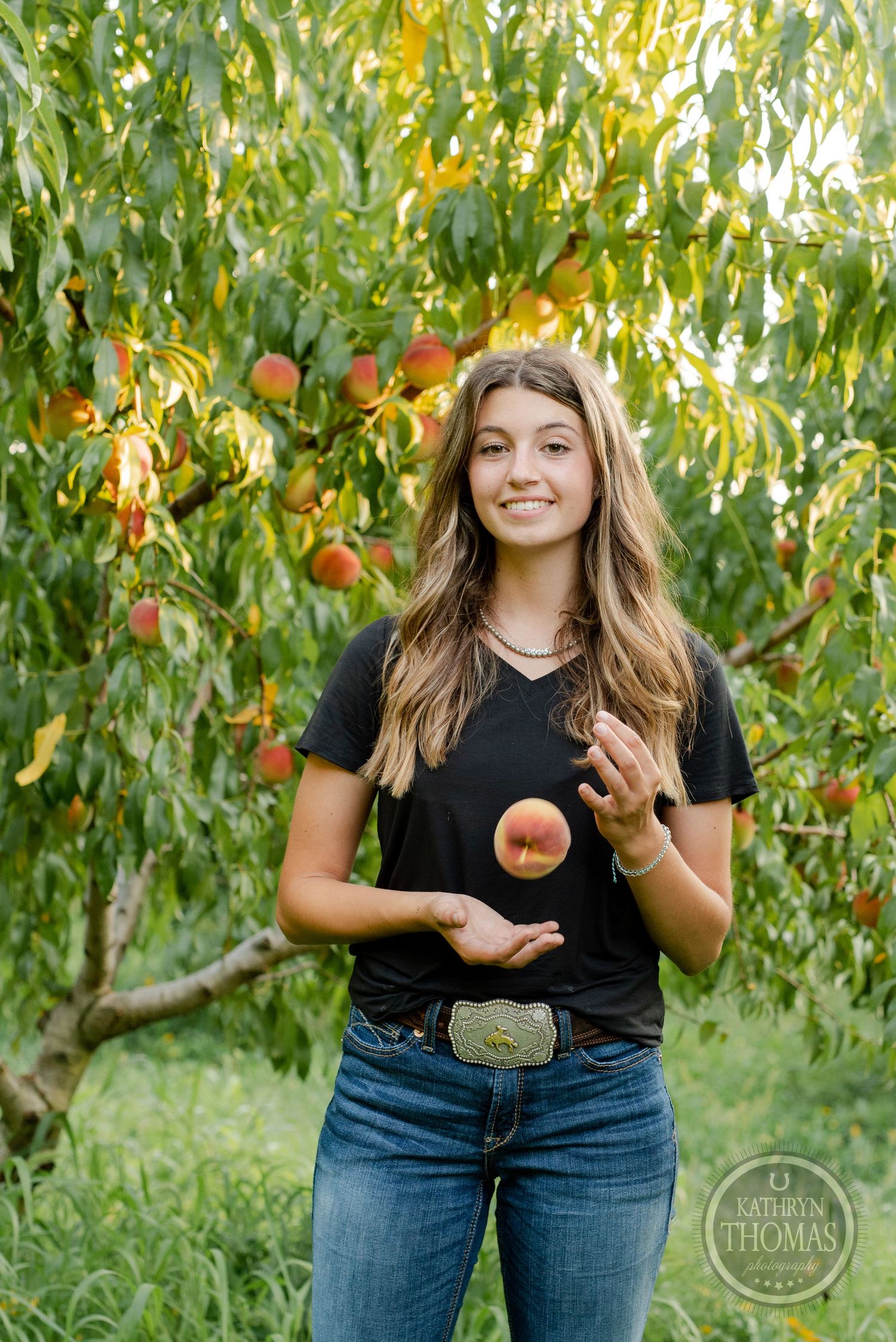 Casual portrait in a peach orchard showing someone tossing fruit while wearing a black t-shirt and jeans.