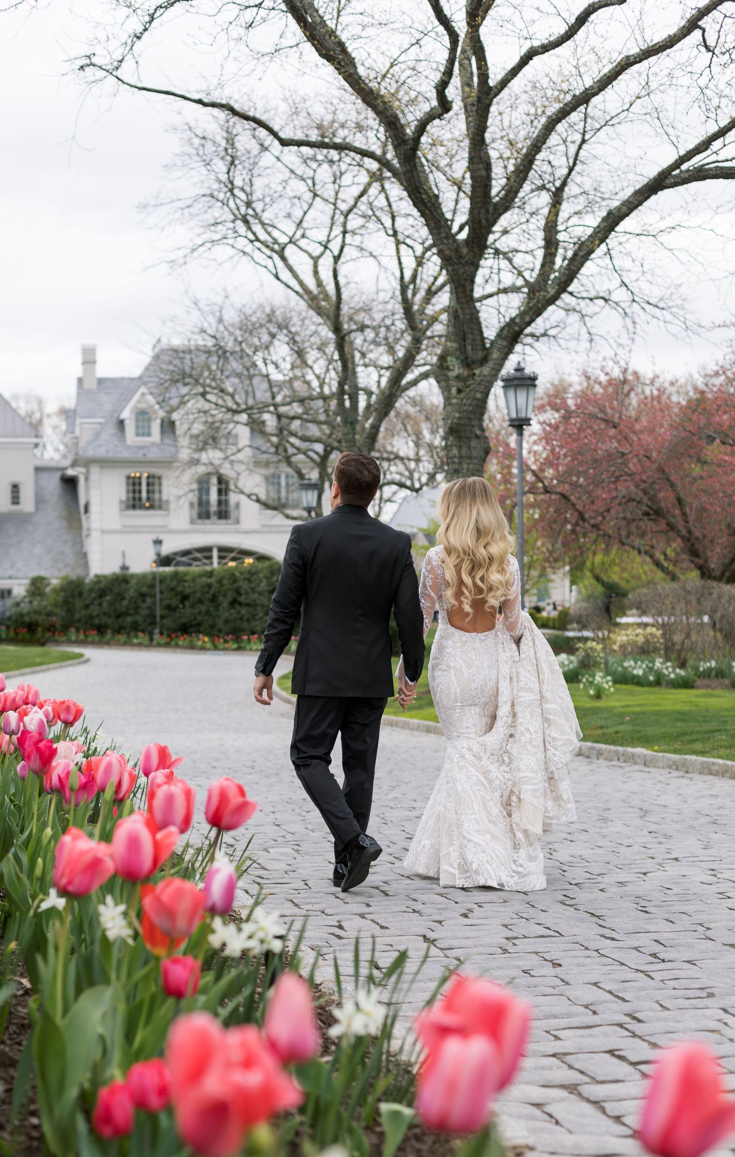 A couple walks along a garden path with blooming pink tulips and a grand house in the background.