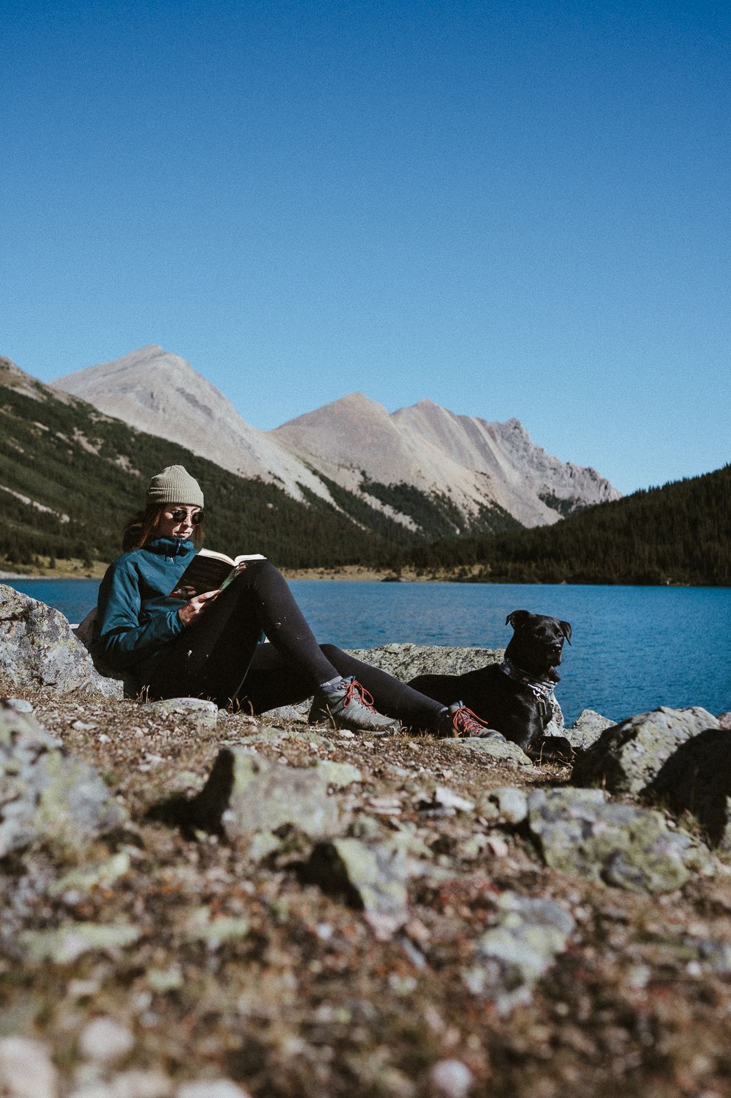 Outdoor enthusiast relaxes by alpine lake with mountain peaks and dog companion on sunny day.