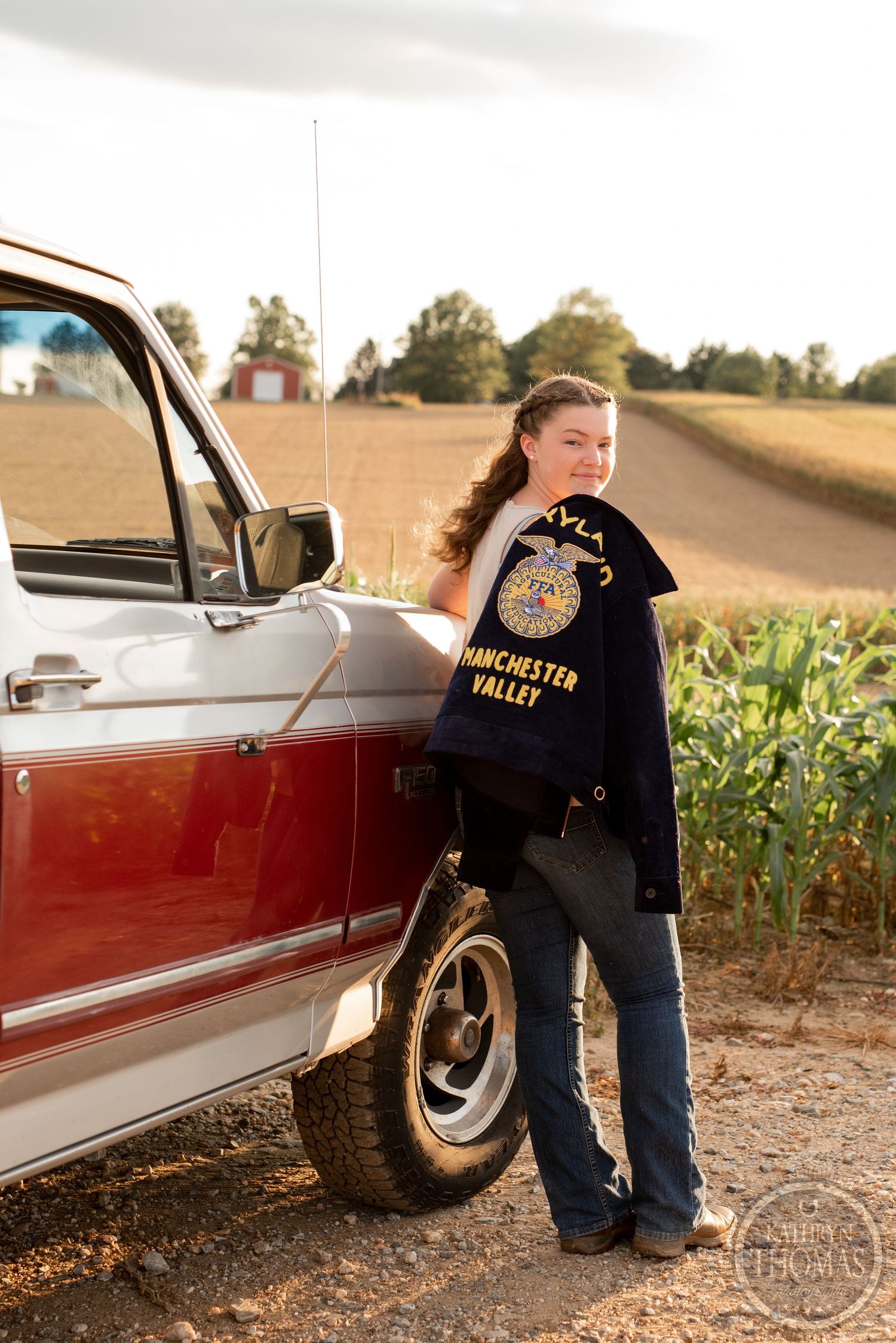 Leaning against a classic red pickup truck while wearing an FFA jacket near cornfields.