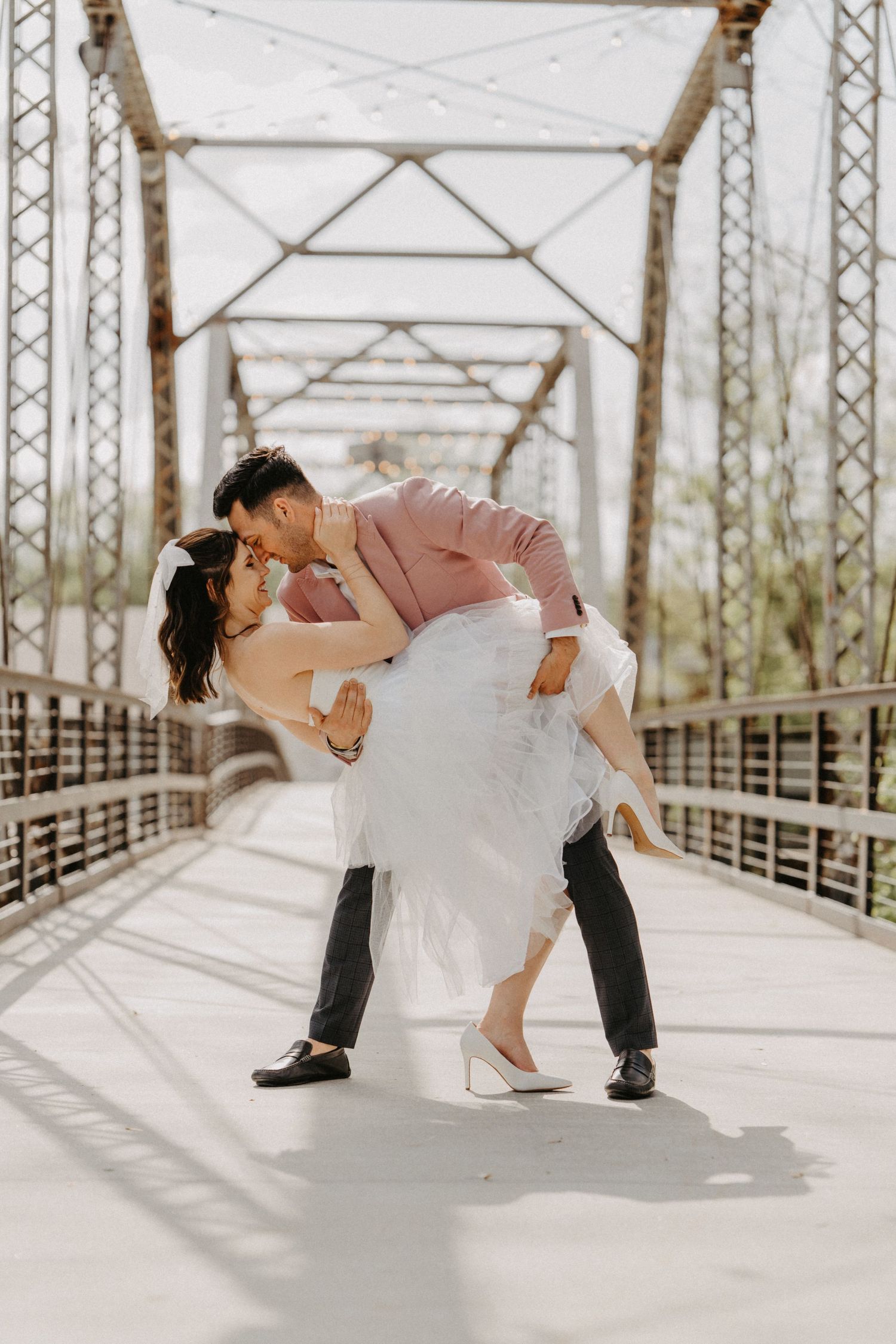Groom in pink blazer dips and kisses bride in white gown on sunlit steel truss bridge.