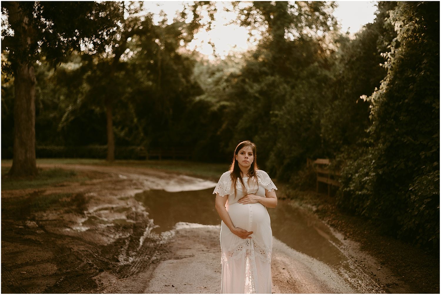 Outdoor maternity photo on dirt path with warm evening light filtering through trees.