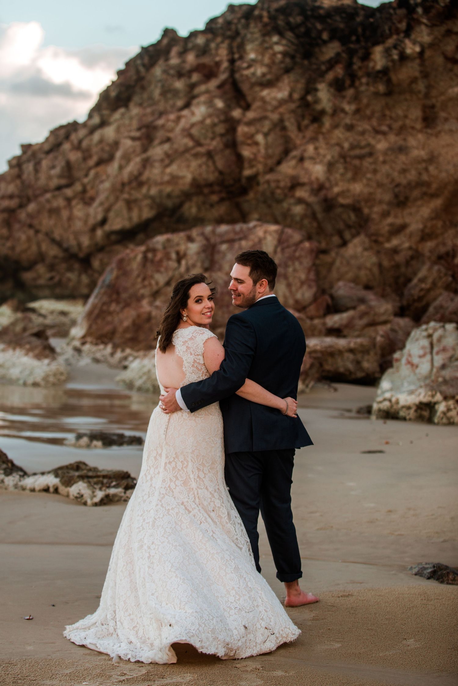 Wedding couple embraces on a beach with dramatic rocky cliffs in the background.