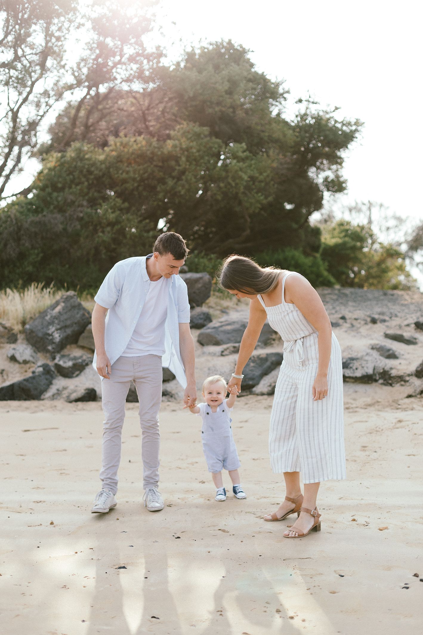 Parents helping toddler take first steps on sandy beach during golden hour.