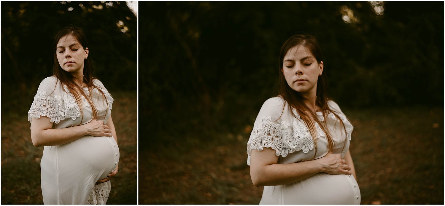 A dreamy maternity photoshoot features a white lace dress against a dark outdoor setting at sunset.