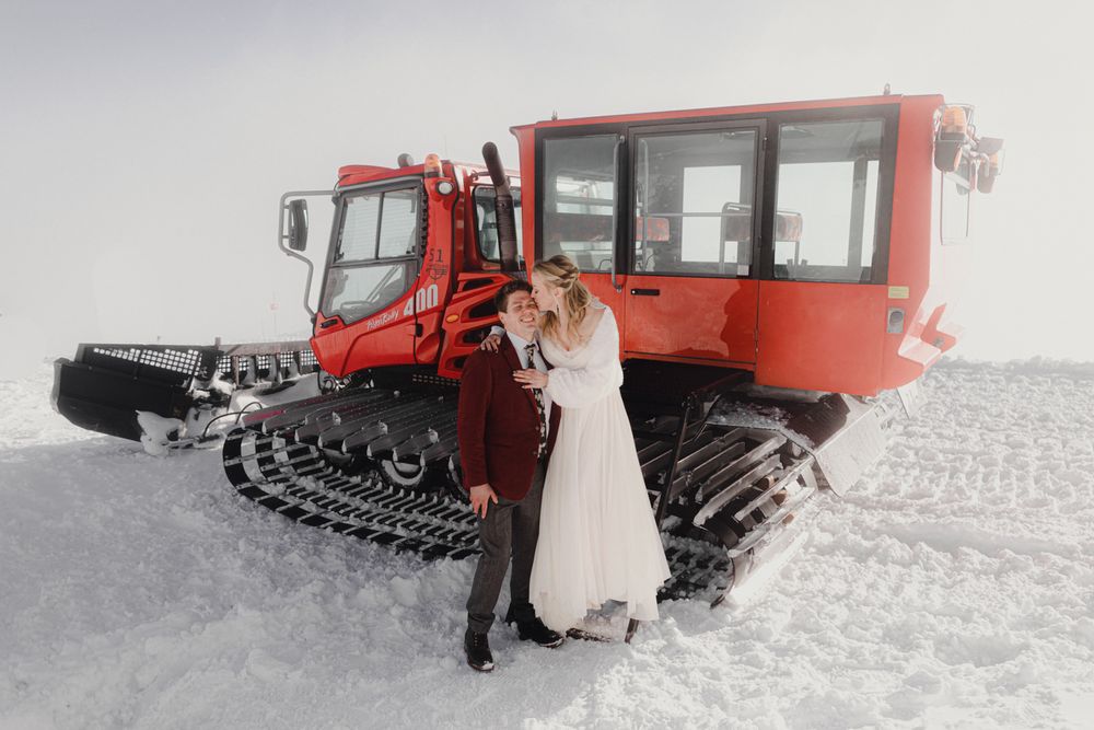 A wedding couple poses in front of a large red snow groomer machine in a snowy landscape.