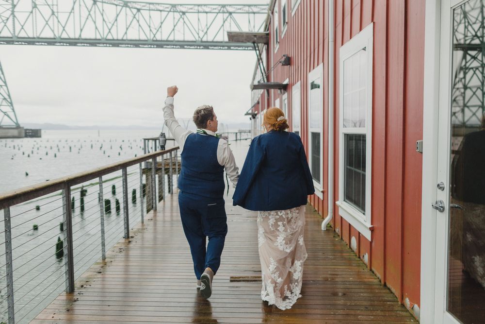 Two people walk along a waterfront boardwalk near an industrial building and bridge.