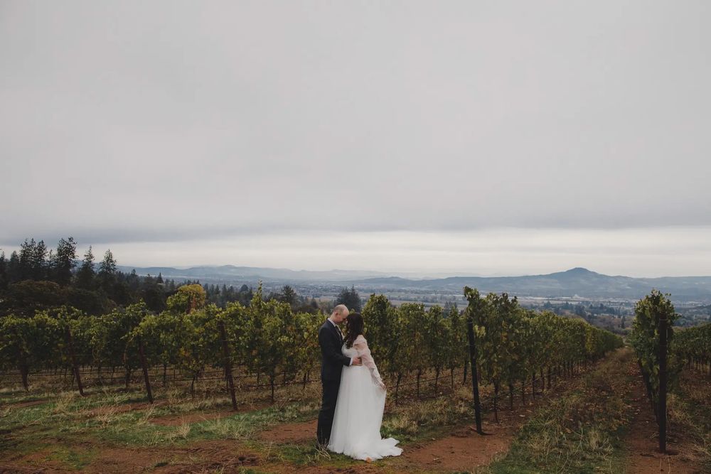 Couple embraces in a scenic vineyard with mountains and overcast sky in the background.