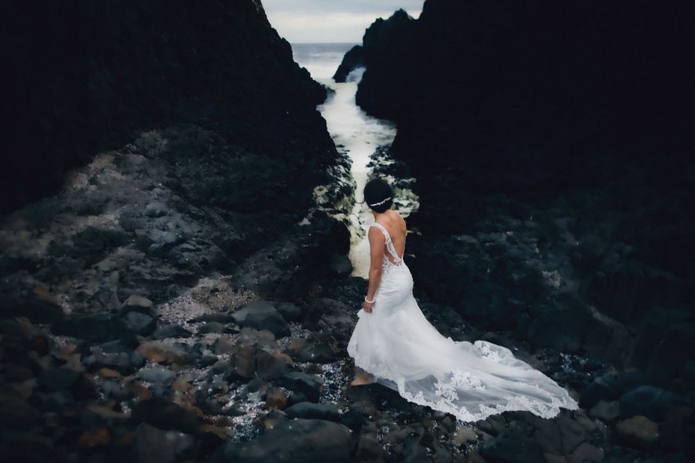 Wedding dress flows dramatically against dark rocky cliffs with ocean waves crashing below.