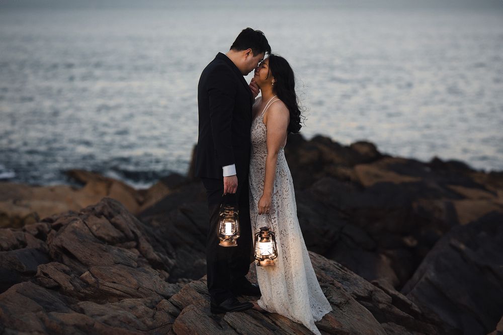 A couple in formal attire holds lanterns on rocky coastal shore at dusk.