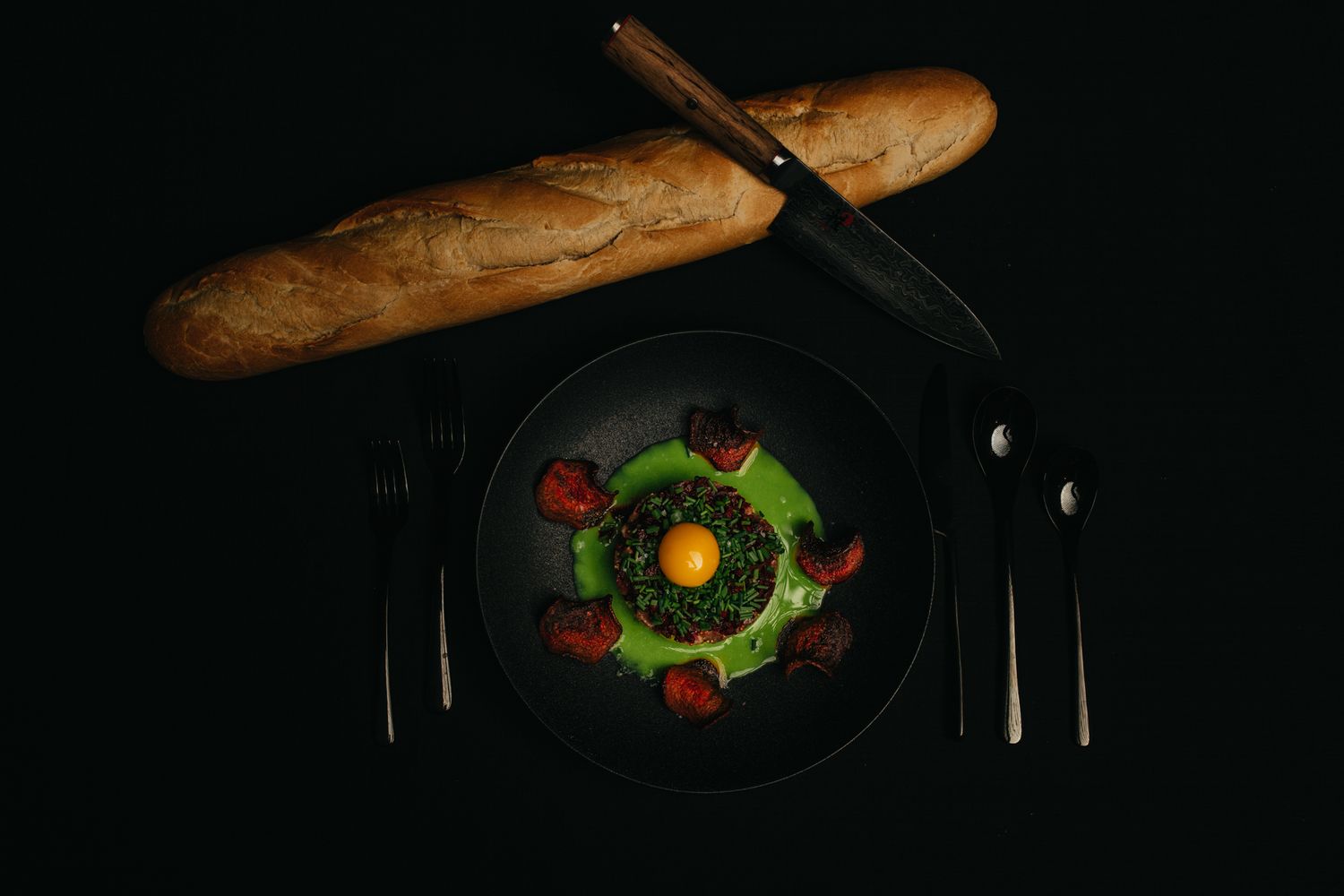 Plating of an elegant dish with colorful vegetables and a crusty baguette on a dark plate.