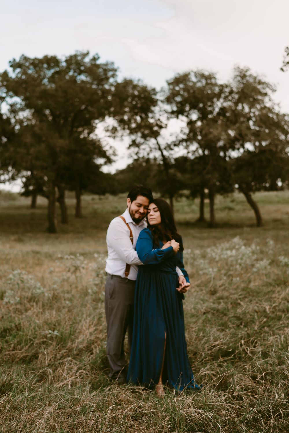 Couple shares an intimate moment in a rustic field setting with scattered trees in the background.