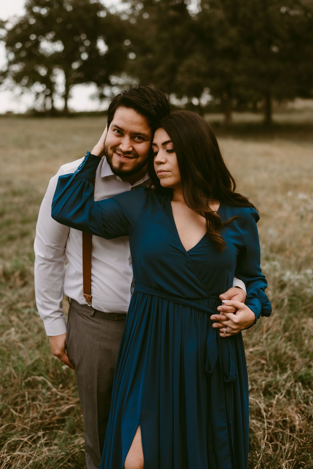 Tender embrace between couple in field wearing navy dress and white shirt with suspenders.