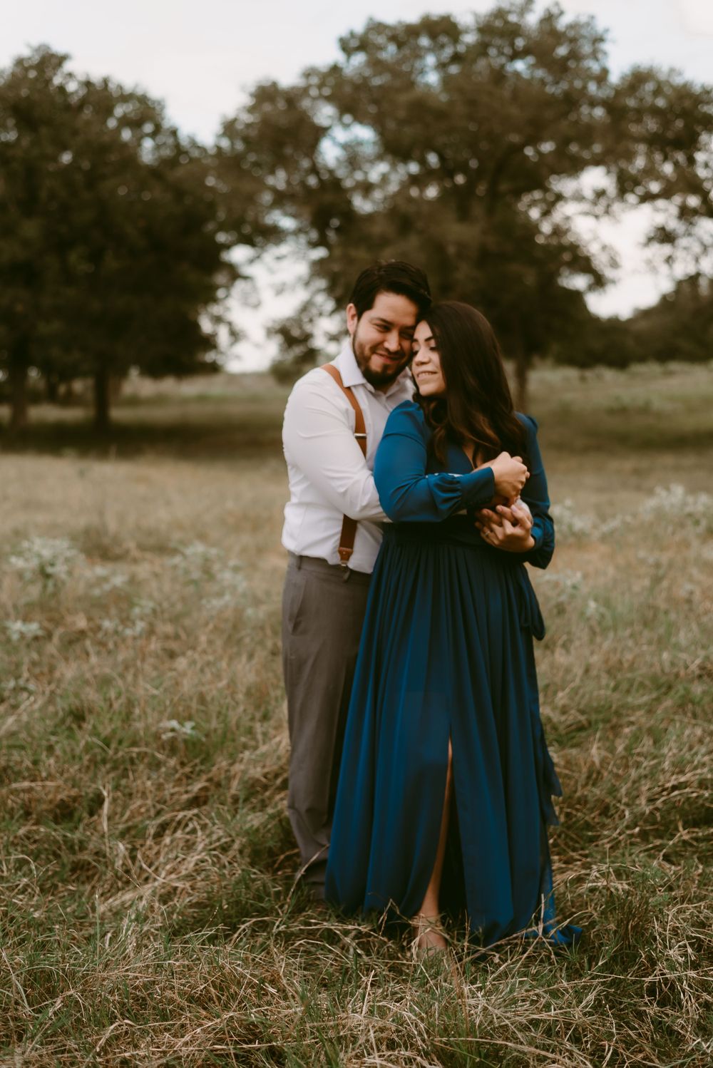 A romantic couple embraces in a field with trees at sunset, wearing formal attire with a navy blue dress and suspenders.