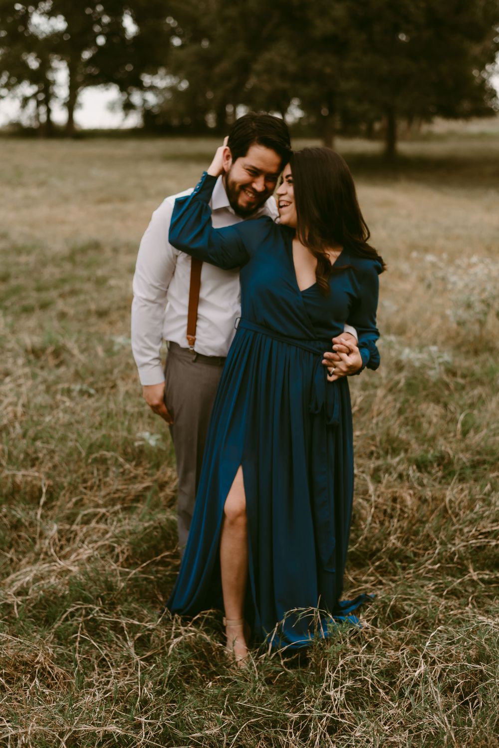 Joyful couple embracing in field wearing navy dress and formal attire with suspenders.