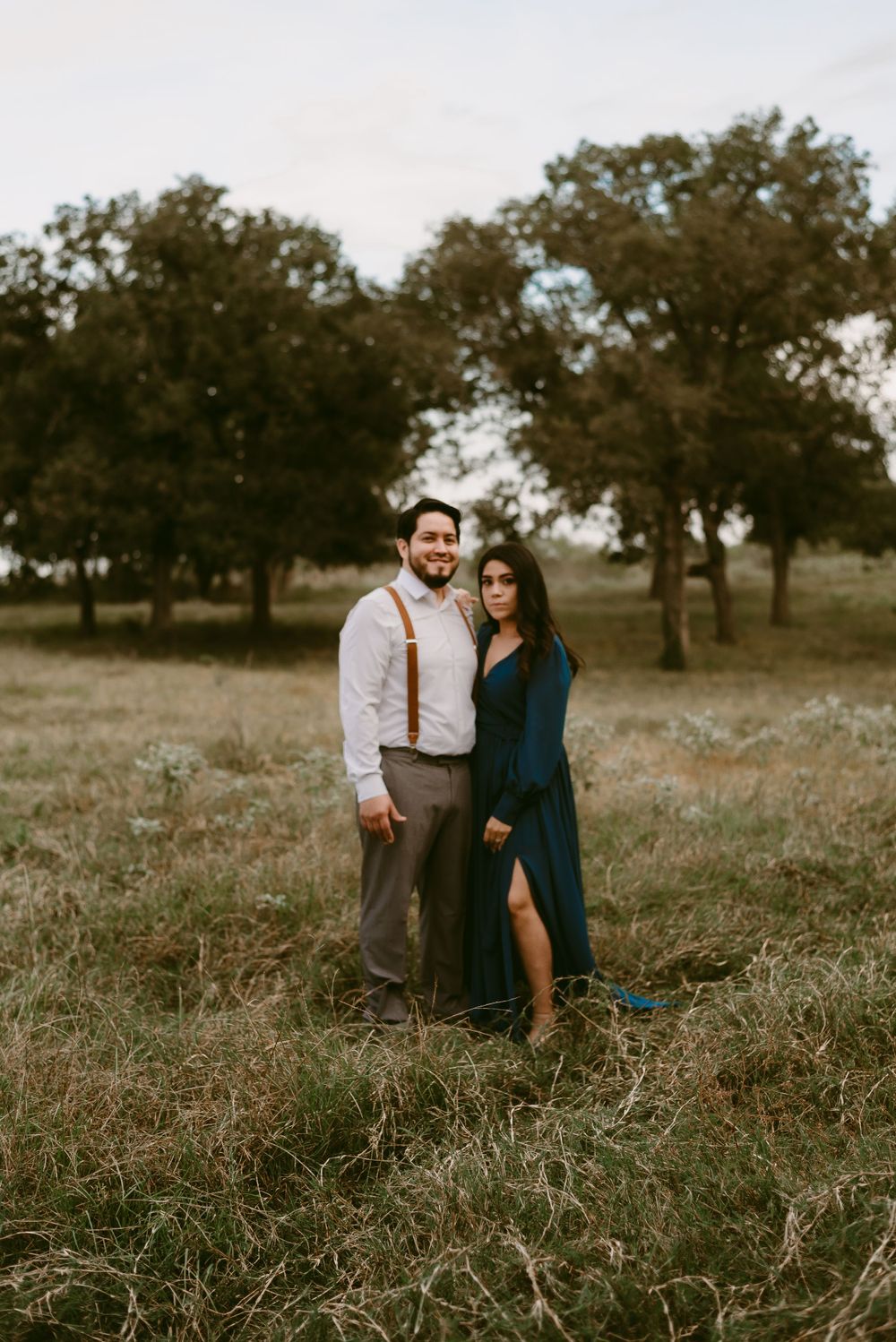 Couple poses in rustic field with oak trees, wearing navy dress and suspenders outfit.