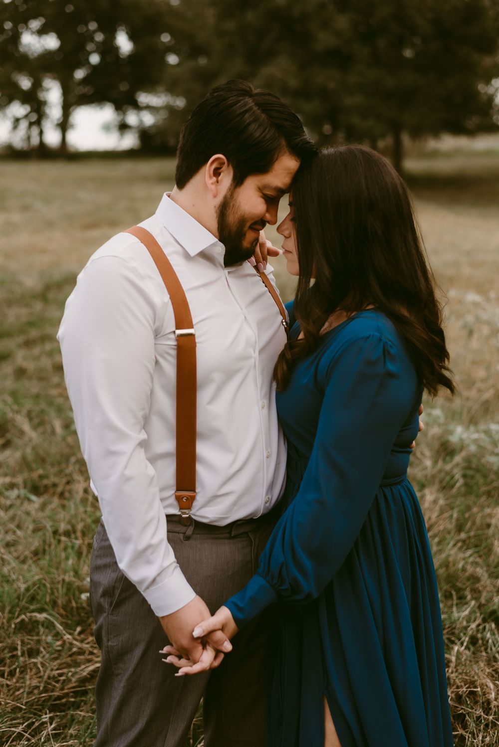 Romantic couple holding hands in field wearing navy dress and white shirt with brown suspenders.