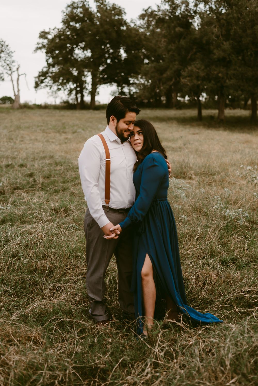 Couple shares tender moment in grassy field wearing formal attire with navy dress and suspenders.