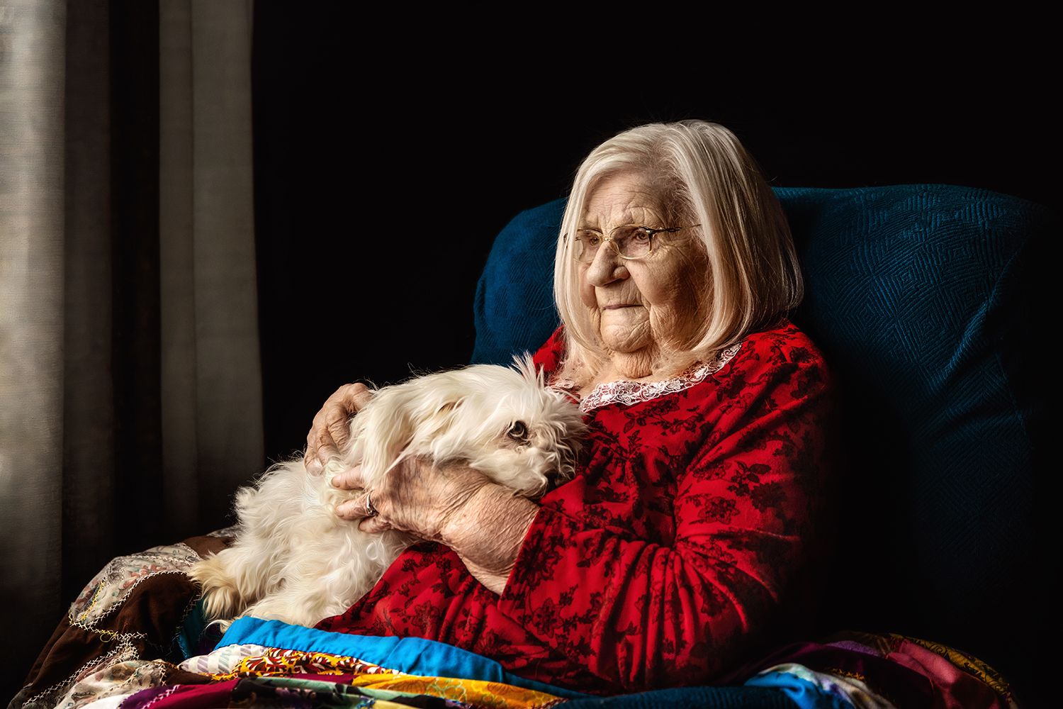 Woman in red sweater cuddling with white dog in blue chair by window.