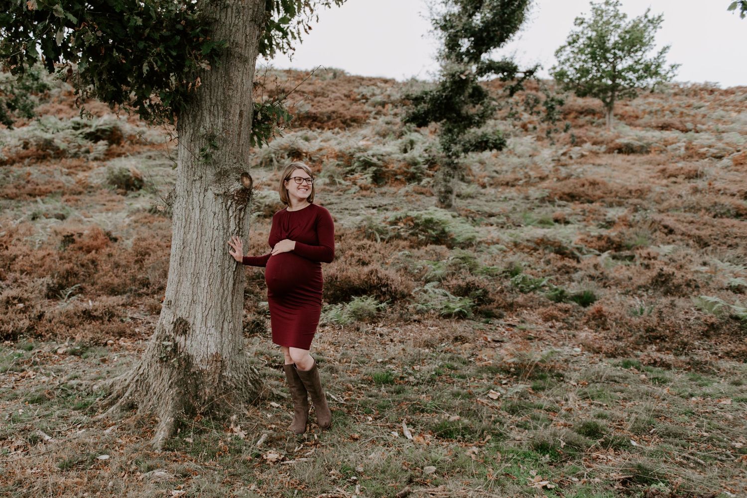 a pregnant lady is stood beside a tree holding her bump, she is looking out into the distance.