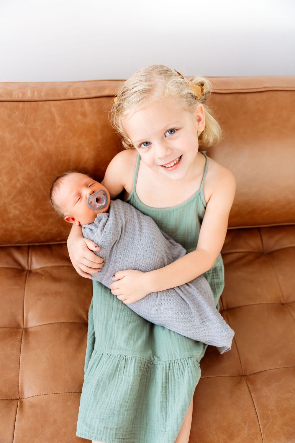 Two siblings share a tender moment on a brown leather couch, with the older sister holding a newborn baby.