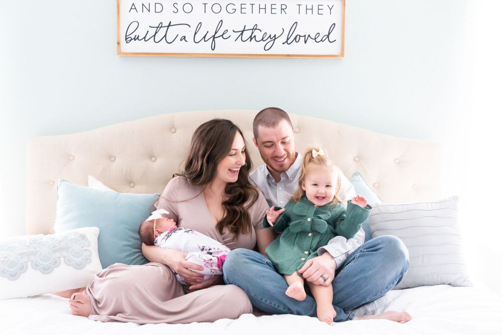 Family relaxing together on bed with light blue pillows beneath wooden wall sign.