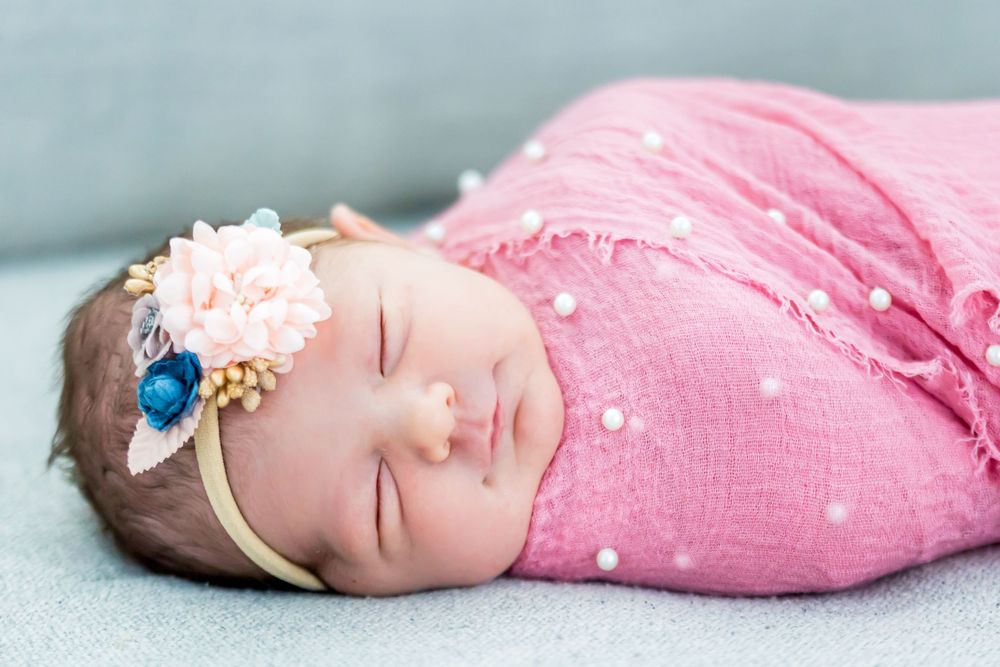 Sleeping baby wearing a floral headband wrapped in pink swaddle on blue background.