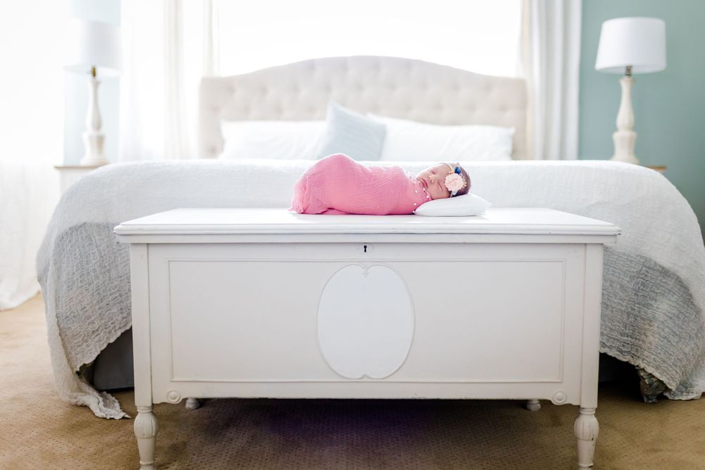 Baby in pink sleeps peacefully on a white storage bench near a tufted headboard.