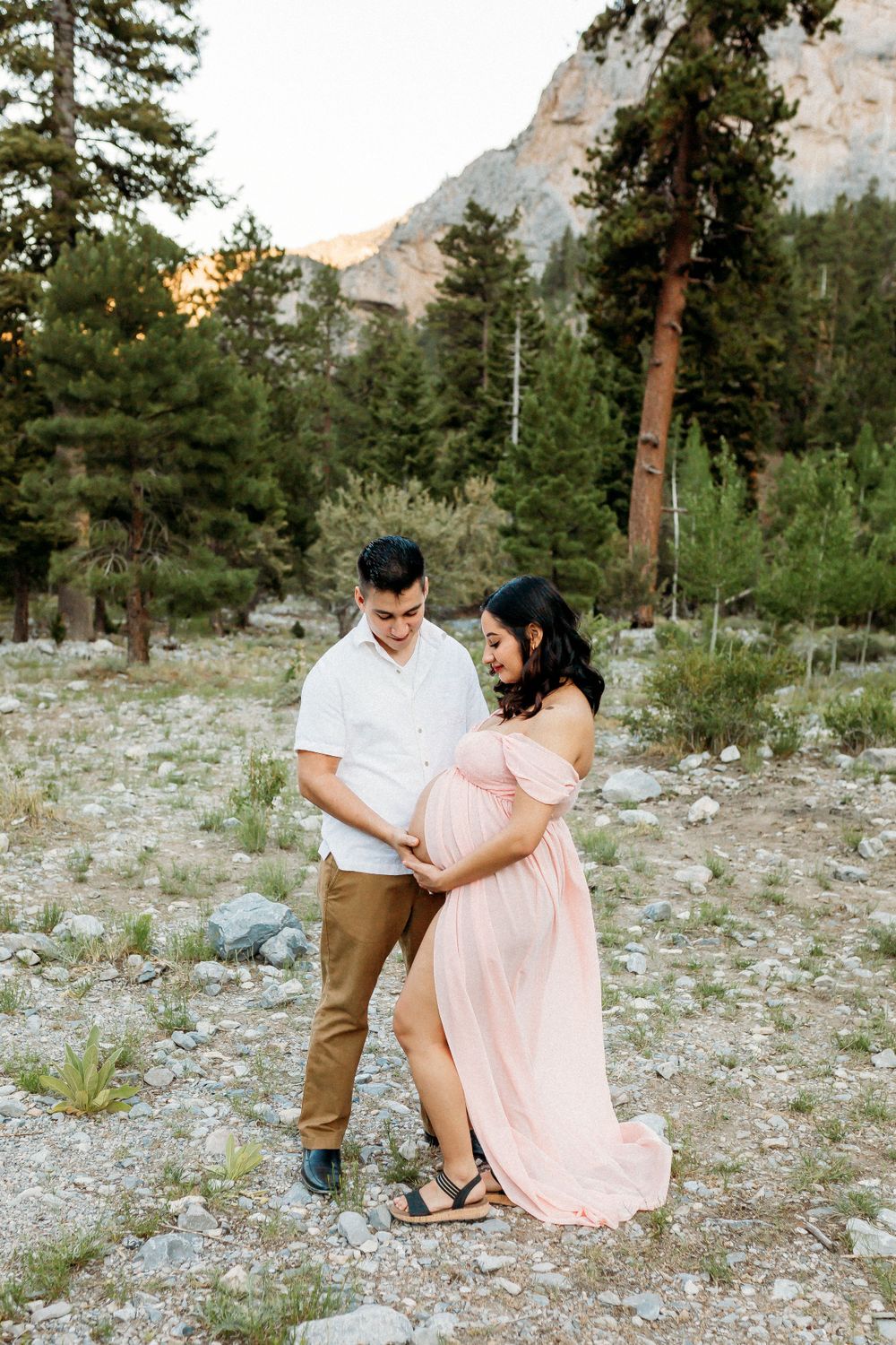 A couple in casual attire poses together in a scenic mountain setting with pine trees.