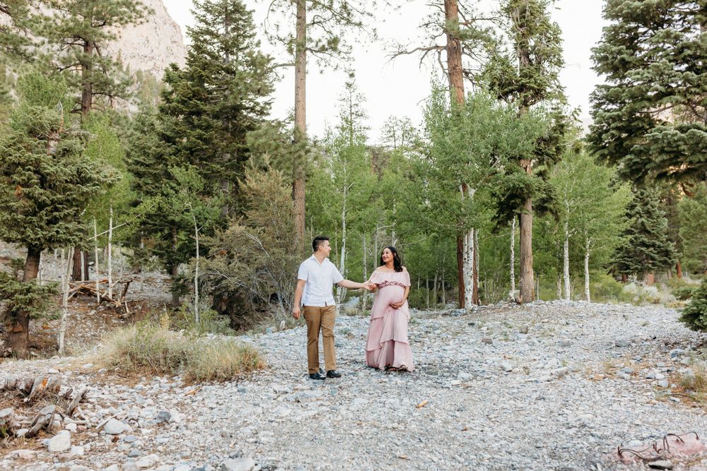 A couple walks hand in hand along a rocky mountain path surrounded by tall pine trees.