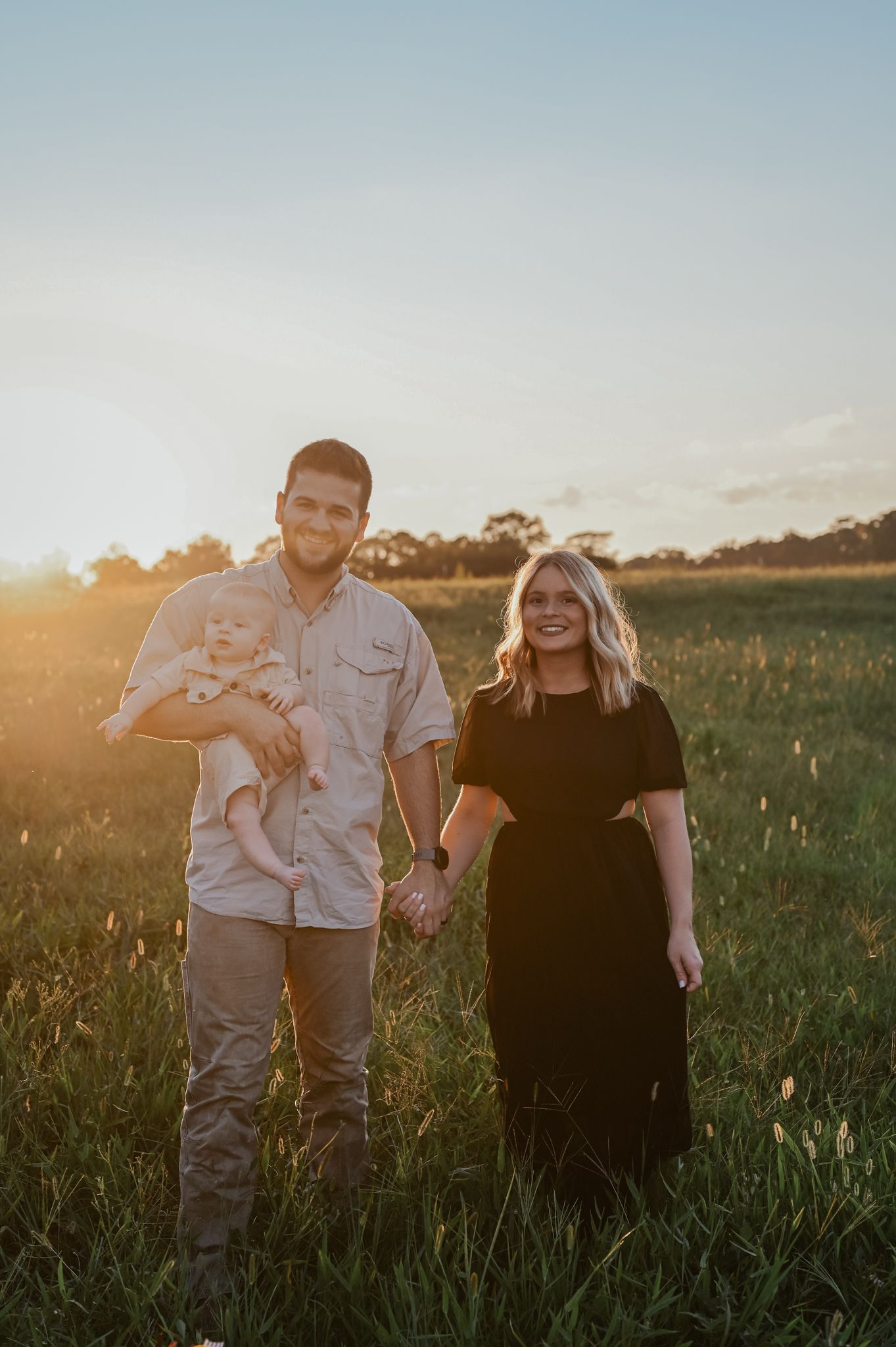 Family of three poses together in a field during golden hour sunset.