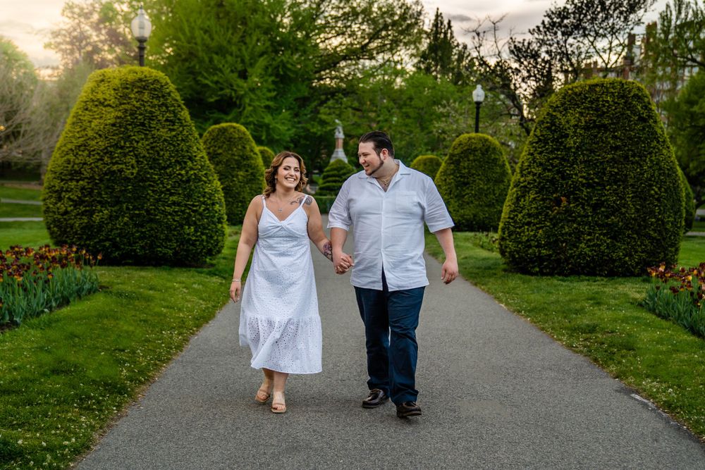 A couple walking hand in hand along a garden path surrounded by manicured topiary shrubs and spring flowers at sunset.