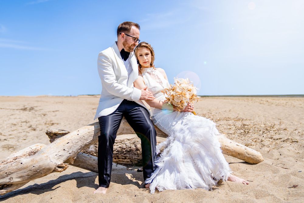 Newlywed couple in white attire posing on driftwood at a sunny beach.