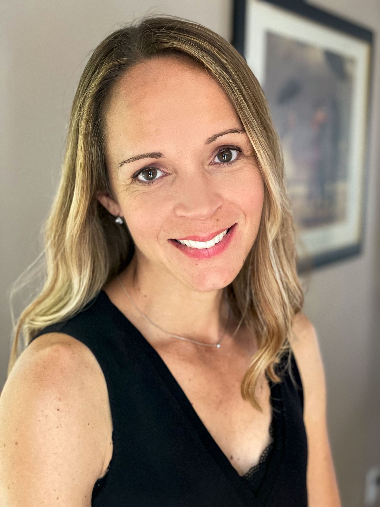 Head shot of owner, in black tank, shoulder length light blonde hair, smiling at camera in her home.