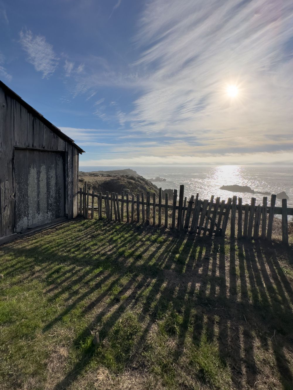 Rustic wooden barn and fence silhouetted against a bright coastal sky with dramatic cloud formations over the ocean. At The Sea Ranch Lodge
