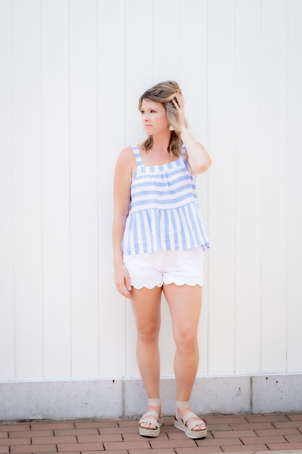 Woman in blue and white striped summer top and white scalloped shorts poses against white wall.
