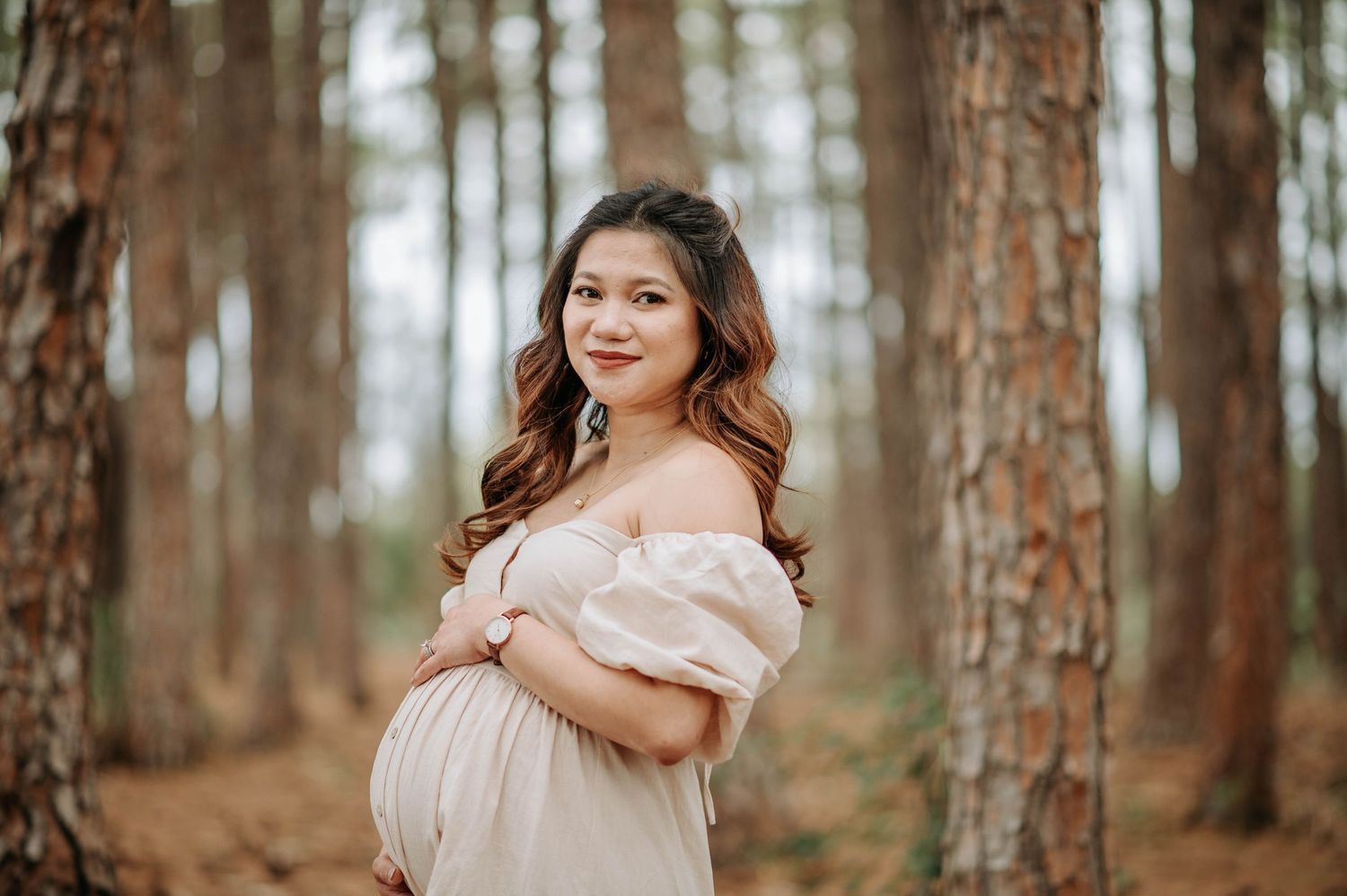 Expecting mother in off-shoulder dress poses gracefully in a pine forest.
