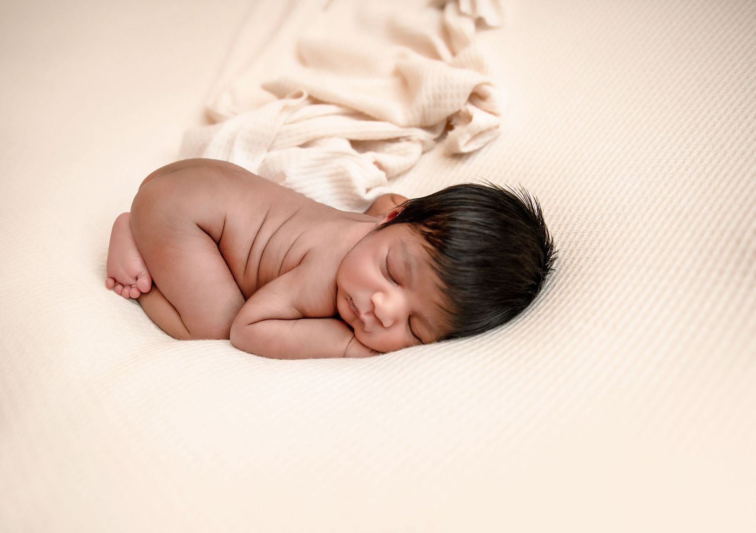 Sleeping newborn baby lying on cream colored textured fabric during professional photo shoot.