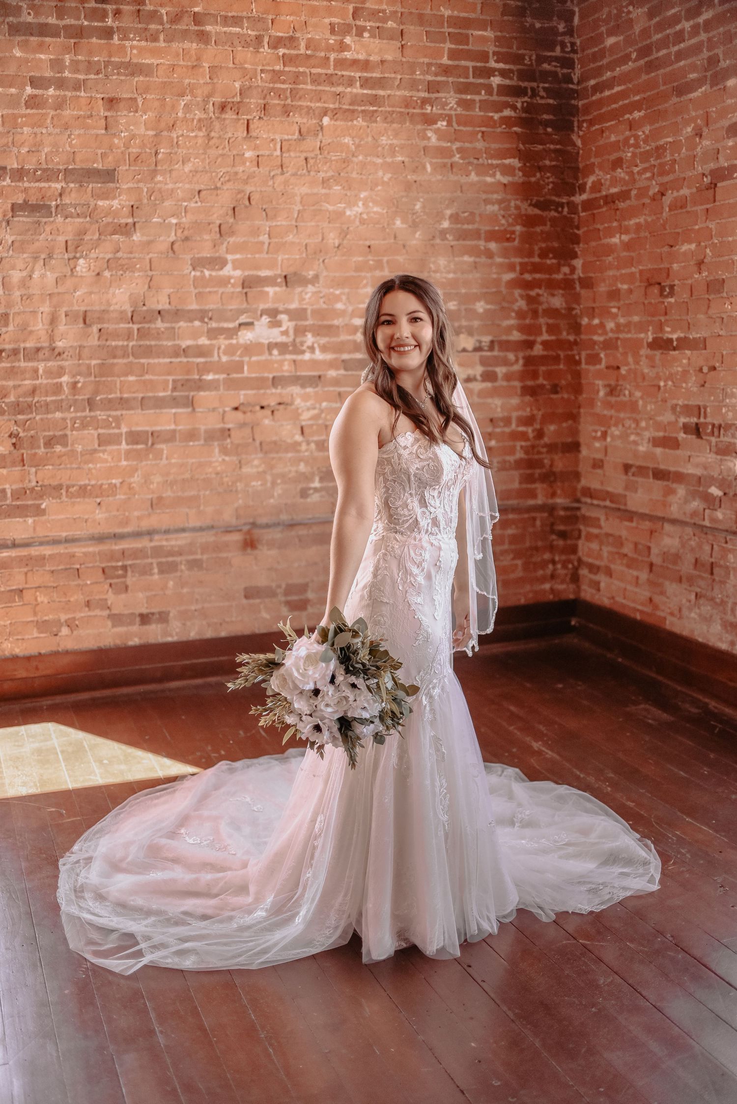 Bride in flowing lace gown posing in rustic brick room with bouquet.