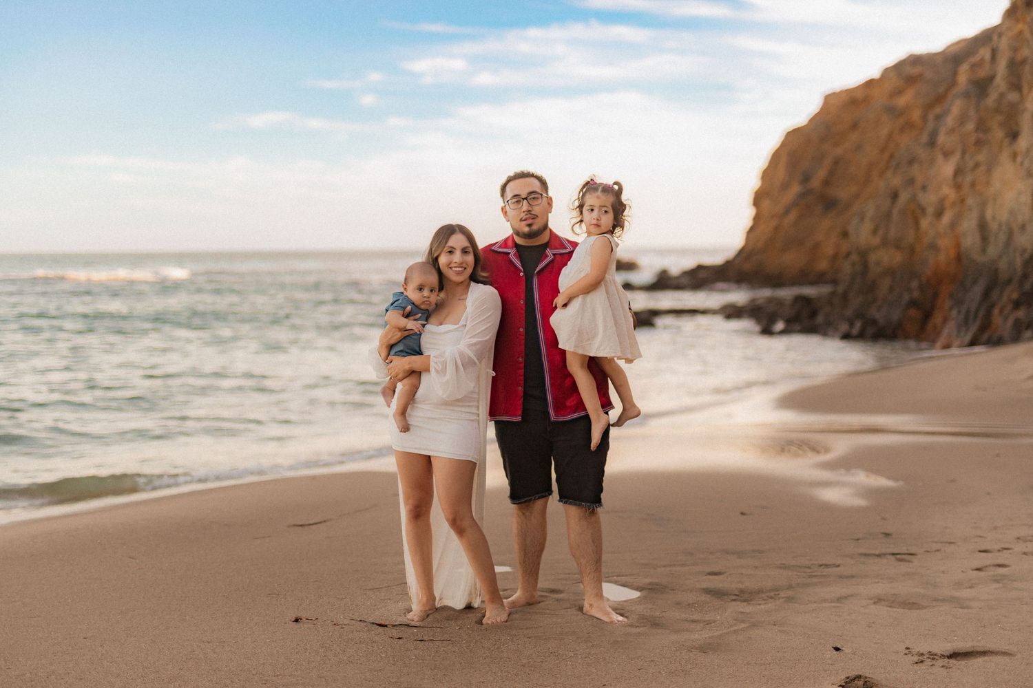 Family of three standing together on a beach with cliffs during sunset.