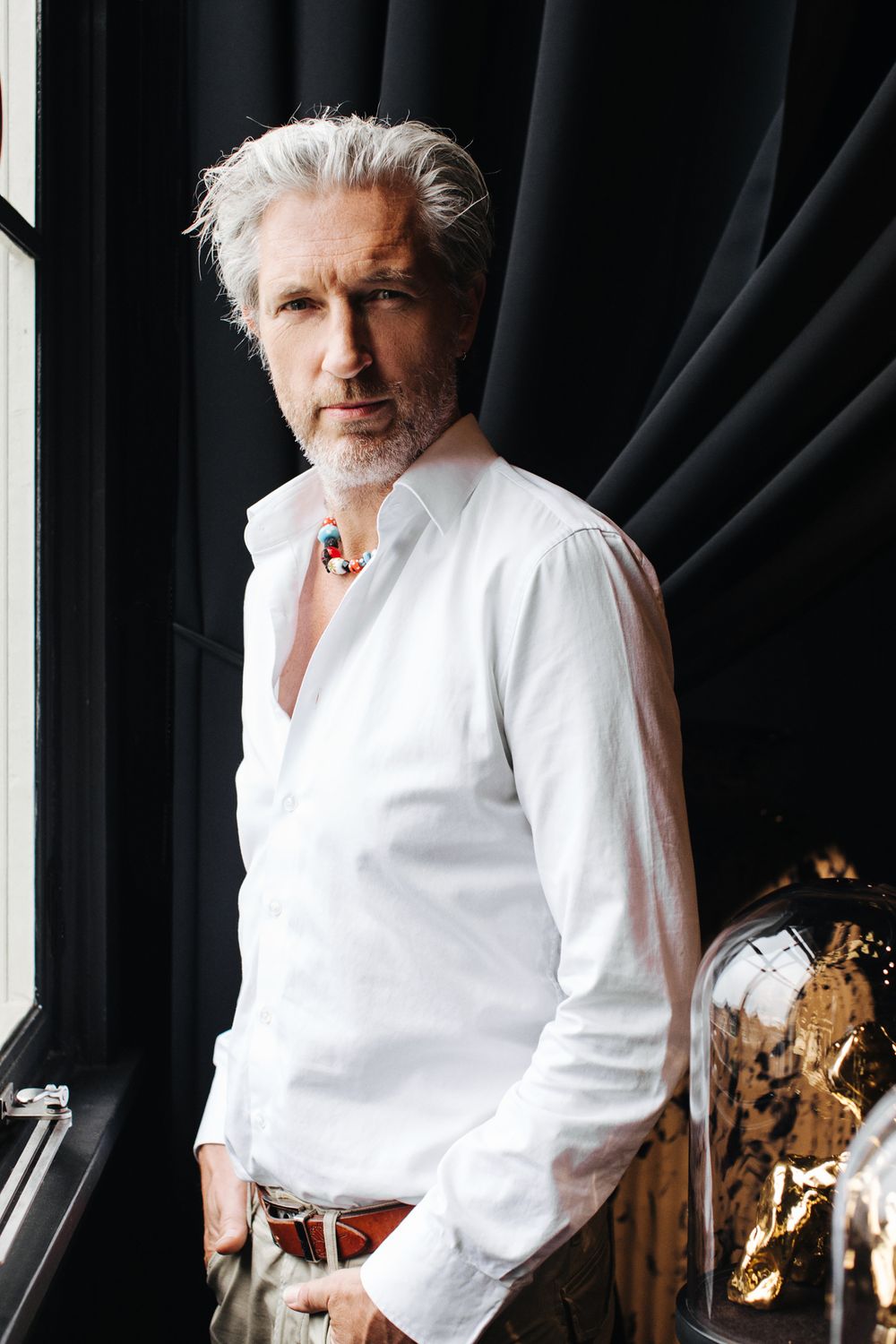 A person in white shirt poses dramatically by a window with dark curtains.