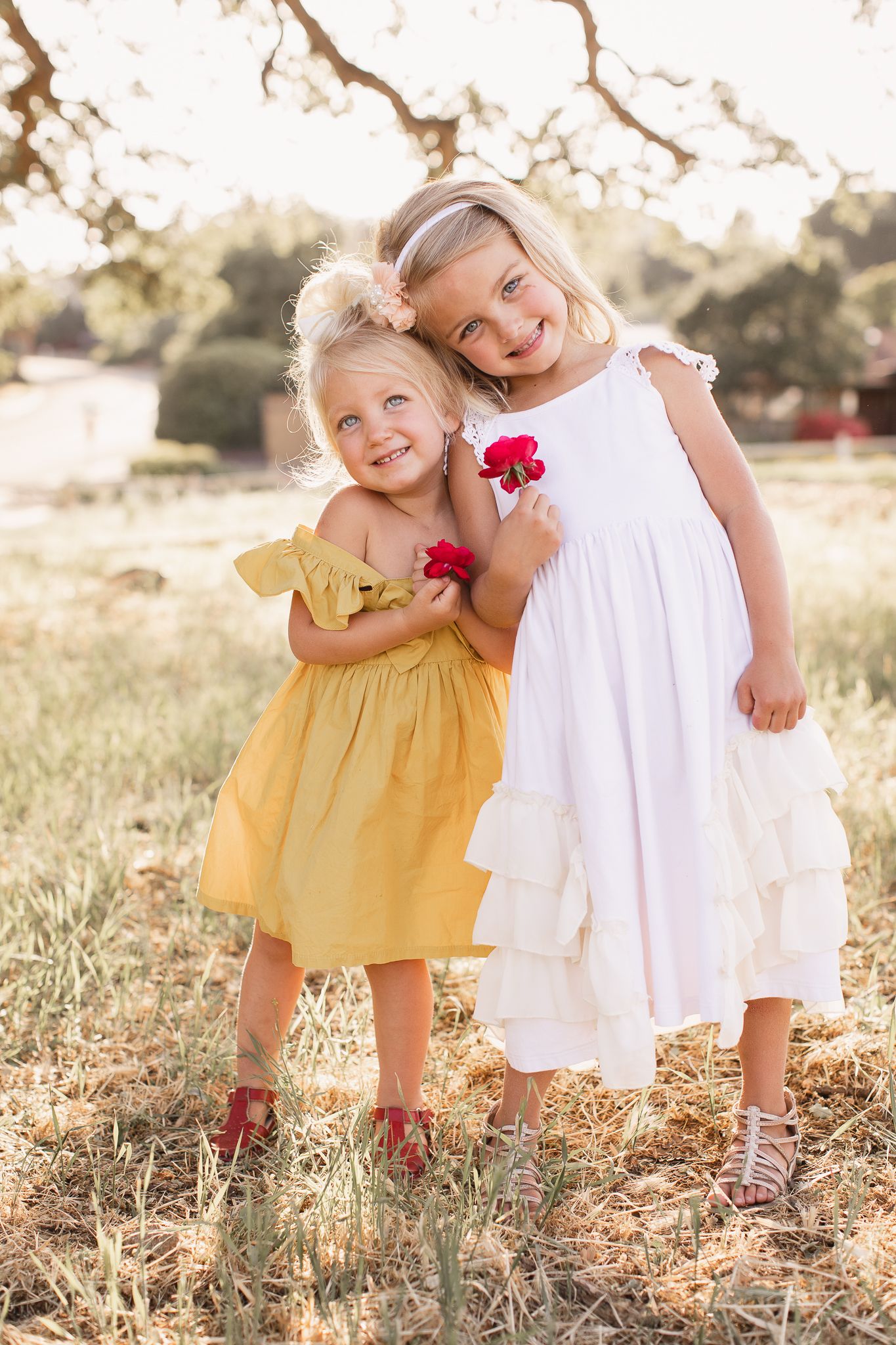 Two young girls in yellow and white dresses hugging in sunny field.