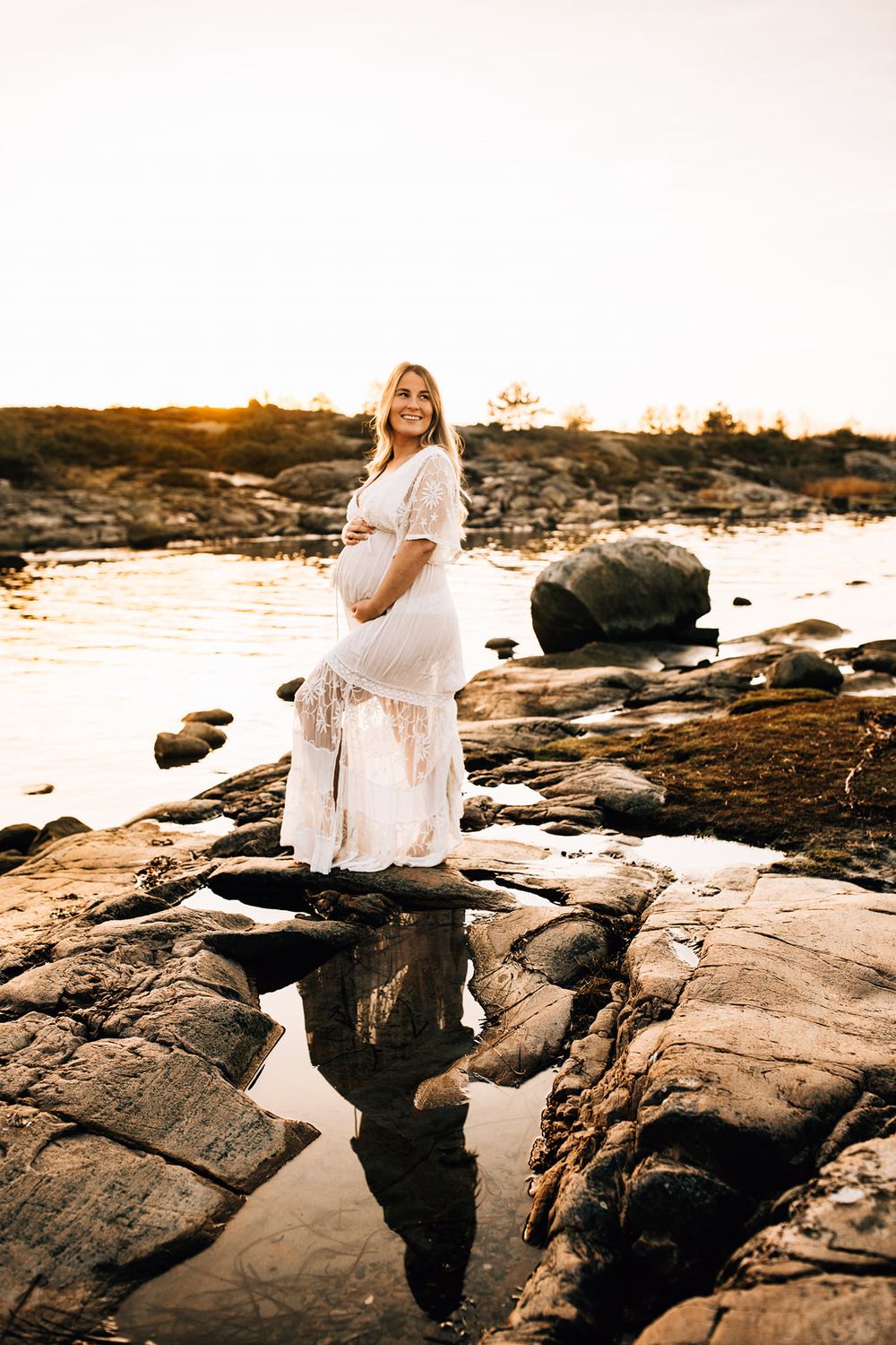Expectant mother in white dress standing on rocky coastline during golden hour.
