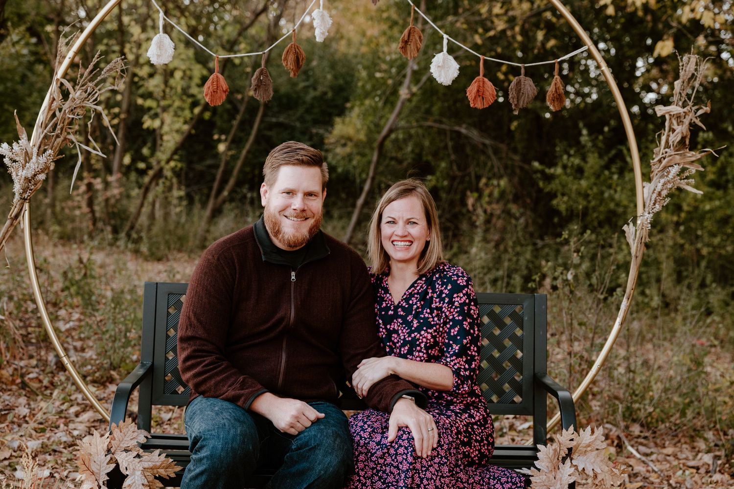 A couple sits on a bench under an arch decorated with string lights and pumpkins.