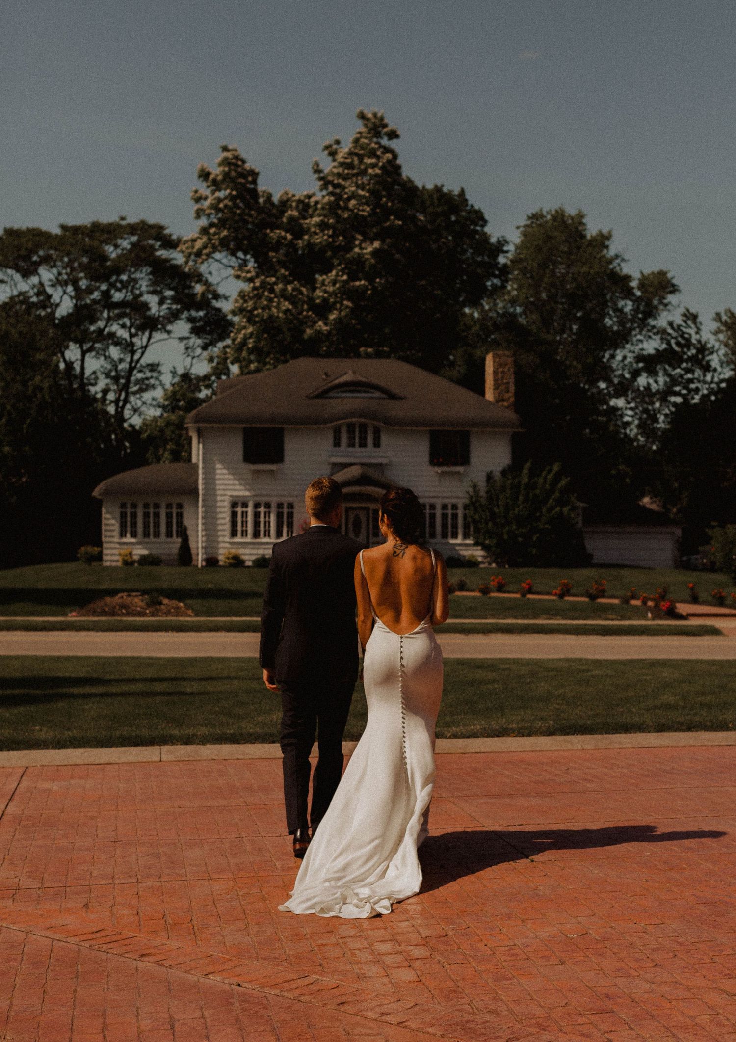 Couple in wedding attire standing in front of an elegant white house with beautiful landscaping.
