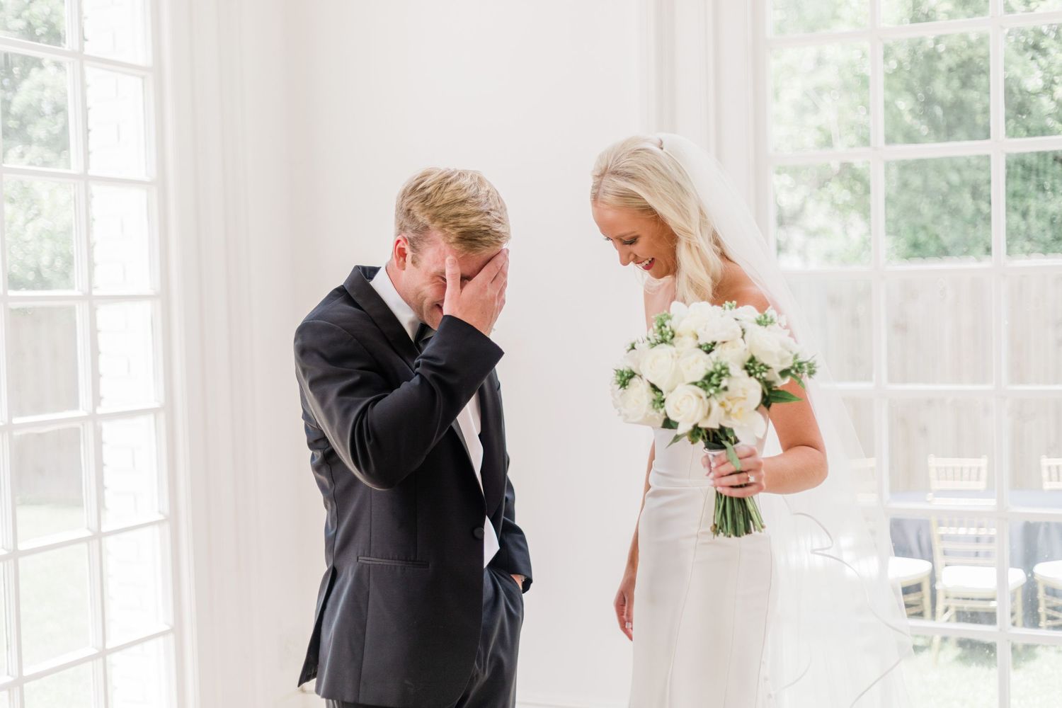 Touching first look moment with white floral bouquet in sunlit room.